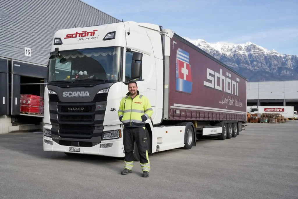 Chauffeur SPL suisse devant un camion Scania blanc et remorque Schöni porte-drapeau suisse, paysage montagneux en arrière-plan. Chauffeur en gilet haute visibilite, poste près du quai de chargement.