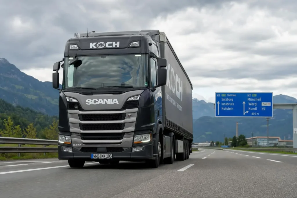 Voiture semitraffic : un grand camion Scania noir sur autoroute européenne, paysage montagneux à l’arrière et panneau routier bleu indiquant plusieurs destinations. Intègre le thème: réglementation transport international de marchandises.